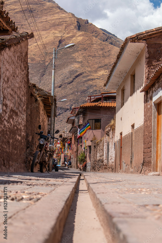 Typical streets in Pisac, with adobo houses, wooden doors, vintage motorcycle and colorful aboriginal flag, with view to the mountains, Peru. 