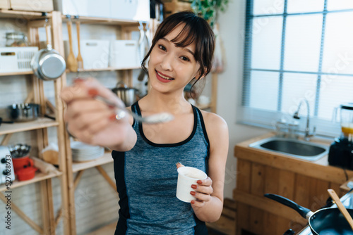 selective focus cute asian girl smiling at the camera in sportswear is feeding her boyfriend yogurt with a spoon at a cozy bright home kitchen background.
