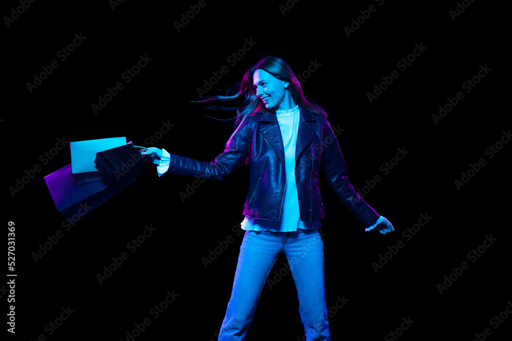 Fototapeta premium Portrait of young excited woman with shopping bags isolated over dark background in neon light. Shopping, sales, ad, black friday concept