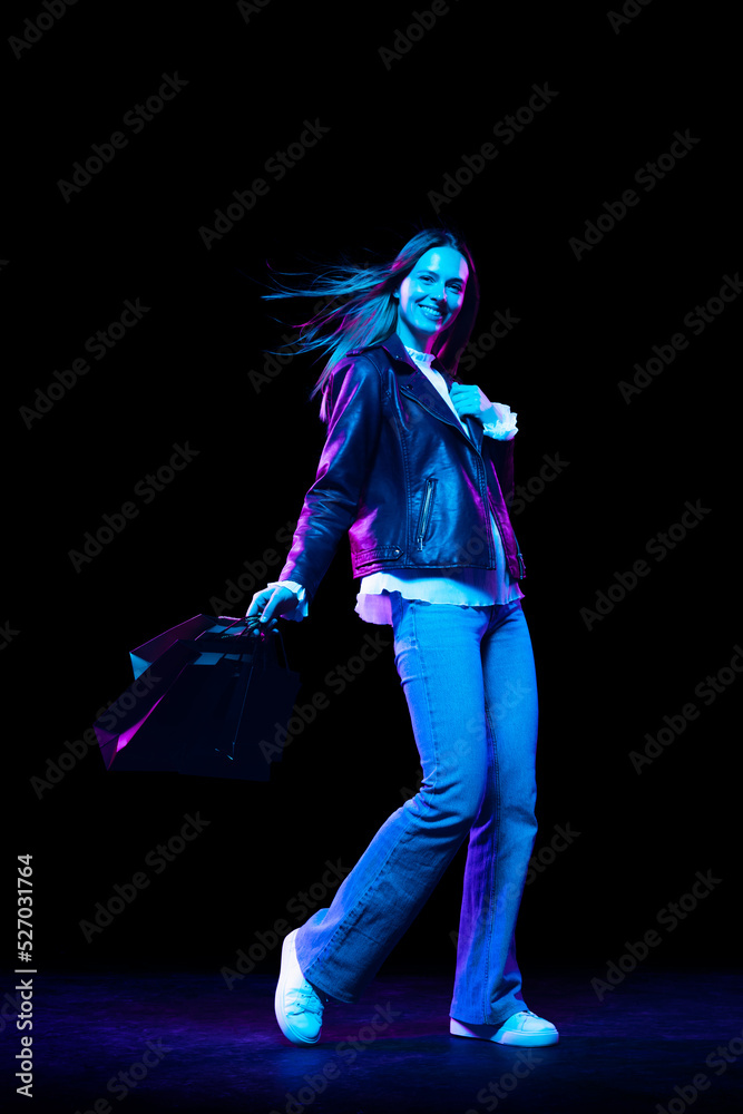 Young fashionable stylish happy woman with shopping bags isolated over dark background in neon light. Shopping, sales, ad, black friday concept