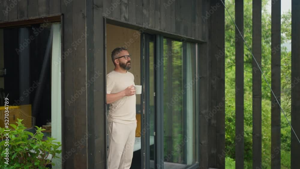 Mature man standing in tiny house terrace in front of beautiful nature with cup of coffee. Morning routine.