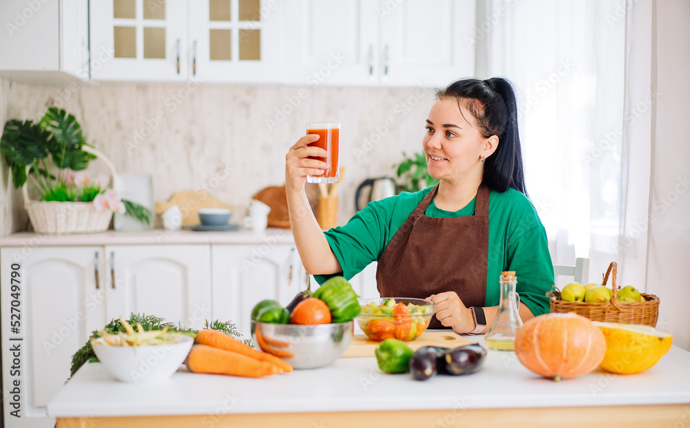 A young brunette woman in an apron is sitting at a table with fresh vegetables, looking at a glass of freshly squeezed homemade juice from fresh vegetables in a white kitchen.