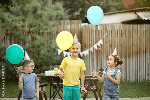 Cute funny kids celebrating birthday with family or friends in a backyard. Birthday party. Kid wearing party hat and launch balloons into the sky photo