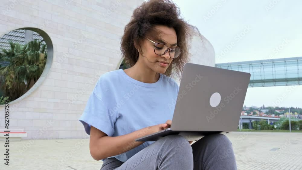 Clever african american young woman with curly hair, female mixed race freelancer, student, with glasses, in a blue shirt, working in a laptop while sitting outdoors, looking at screen, smiling