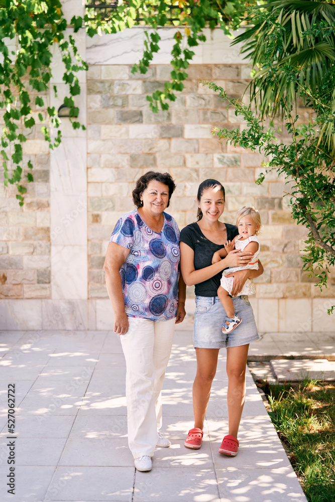 Grandmother stands next to mother holding a little girl in her arms in a garden near a stone wall