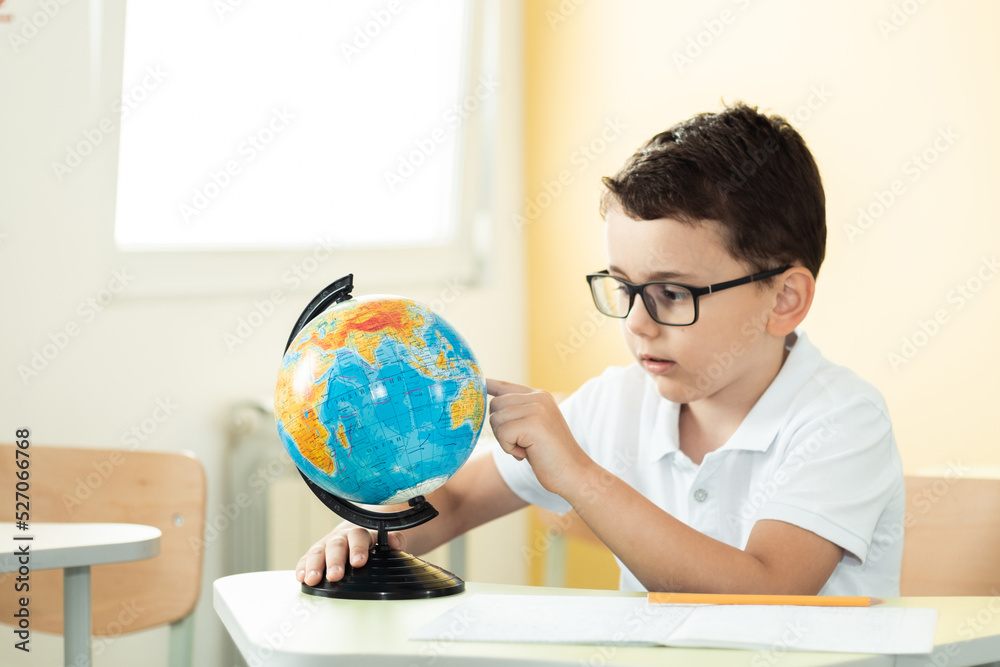 Cute caucasian schoolboy wearing glasses and sits at a desk in the classroom at elementary school. Little boy next the Globe at school. Back to school concept Cute caucasian schoolboy wearing glasses and sits at a desk in the classroom at elementary school. Little boy next the Globe at school. Back to school concept