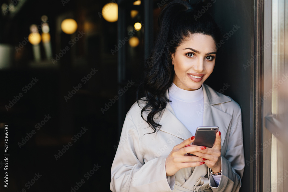 Gorgeous brunette woman with dark eyes, nice make-up and full lips dressed in white raincoat holding modern cell phone surfing social networks online. People, youth, lifestyle, technology concept Gorgeous brunette woman with dark eyes, nice make-up and full lips dressed in white raincoat holding modern cell phone surfing social networks online. People, youth, lifestyle, technology concept