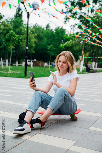 A young beautiful teenage girl is sitting on a skateboard on a beautiful summer day in the park. A girl is talking by video on a mobile phone in the open air. Chat with friends.