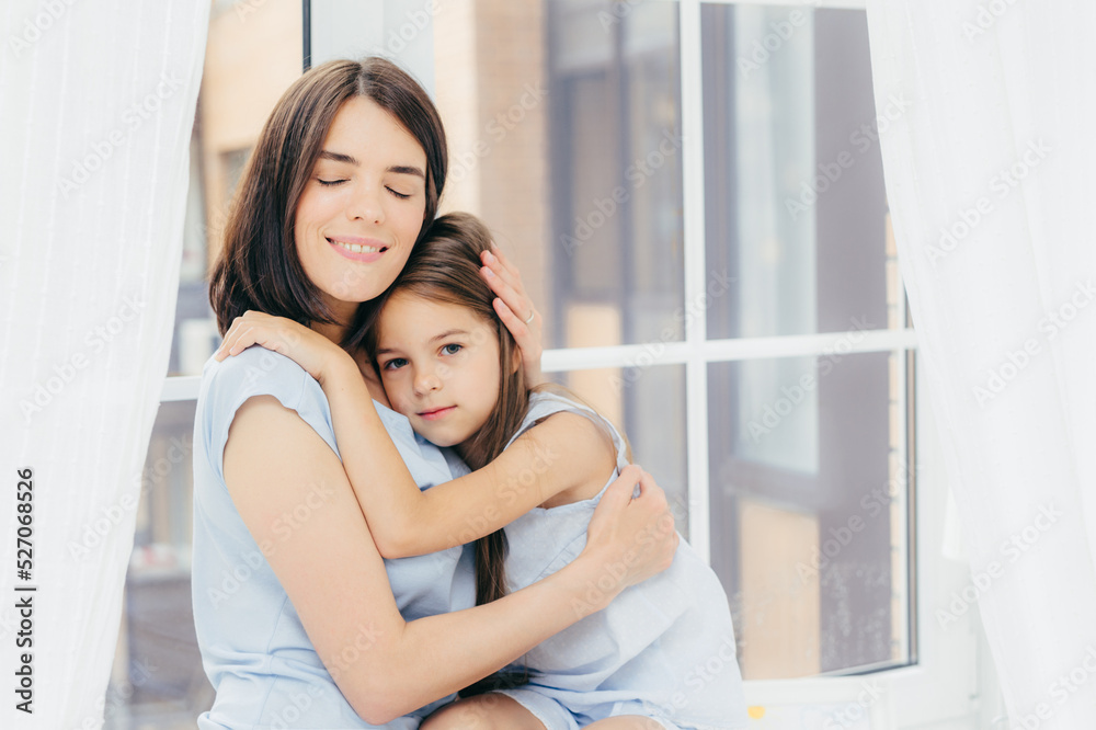 Little female child sits close to her mother, feels support and love of her mum, pose together against window sill background. Delighted mom upbrings small daughter. People, home, family and relations Little female child sits close to her mother, feels support and love of her mum, pose together against window sill background. Delighted mom upbrings small daughter. People, home, family and relations