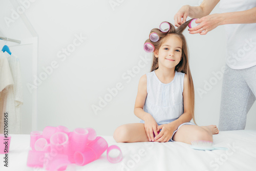 Horizontal shot of cute small daughter sits near her mother who winds curlers, pose against white background, prepares for festive event in kindergarten. Beautiful delighted kid has long hair
