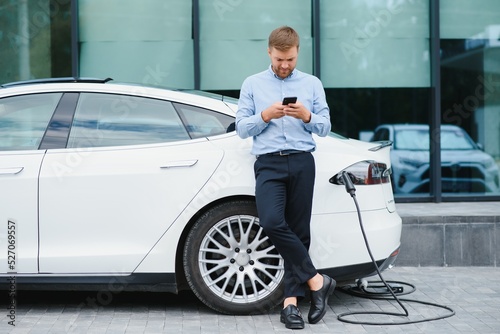Man charging his electric car at charge station and using smartphone © Serhii