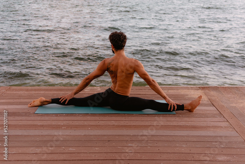 Young beautiful man practicing ashtanga yoga at sunset on the beach. Doing exercises and stretching. Monkey Pose, legs flexibility, split