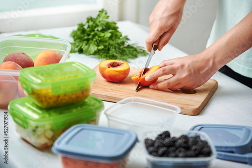 Fototapeta Naklejka Na Ścianę i Meble -  Hands of young female chopping fresh peaches on wooden board while preparing fruits and vegetables for freezing. Plastic containers with raw cut vegetables for freezing on kitchen table.
