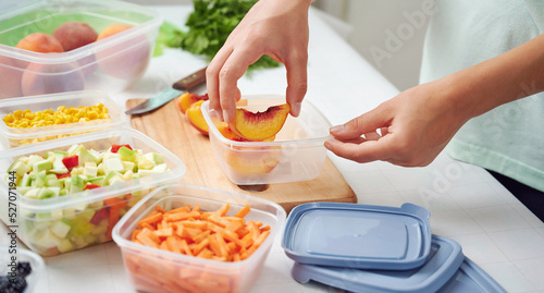 Woman holding plastic container with slices of peaches at table in kitchen. Raw fruits and vegetables for freezing for winter storage in trays