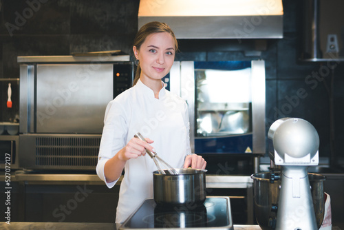 Portrait of confident and smiling young woman chef dressed in white uniform, professional kitchen are on background. Restaurant kitchen