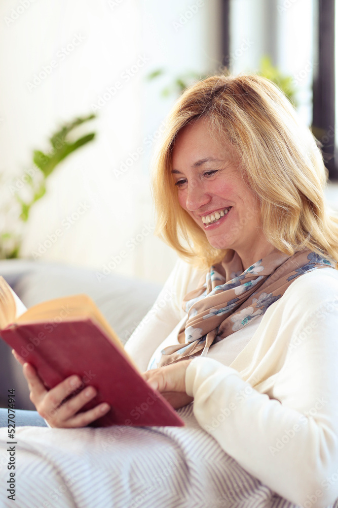 A blonde middle-aged woman sitting in a couch and reading a book at home A blonde middle-aged woman sitting in a couch and reading a book at home