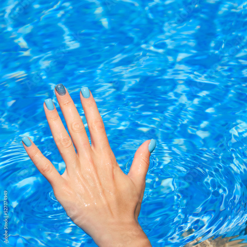 Hand with bright nail polish inside of water. Close-up of female manicured hands. Woman's hand with bright glitter blue fingernails inside of bright blue swimming pool water.