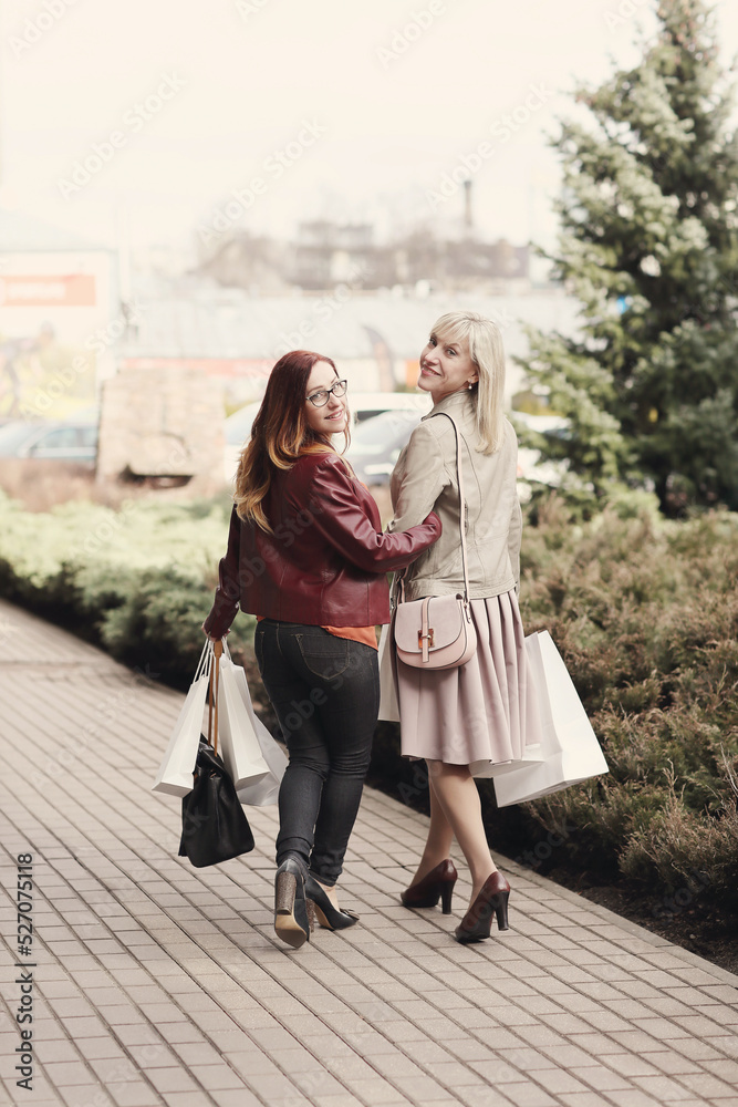Two middle-aged girlfriends holding shopping bags and walking on the street