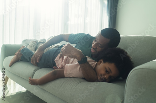 Family spending time at home. Happy African American father and little boy cuddling lying on couch with evening sunbeams shine down in living room at home. Sweet moments of fatherhood concept.