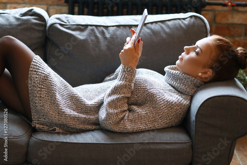 A cute, pretty girl with a bun on her head in a cozy sweater lying with a digital tablet on the sofa photo