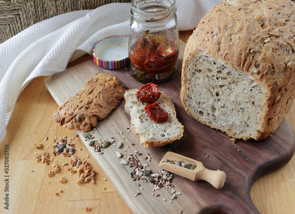 loaf of homemade whole grain bread and a cut slice of bread on a wooden cutting board. A mixture of seeds and whole grains. A jar of sun-dried tomatoes on a crust of fresh bread. Healthy food concept