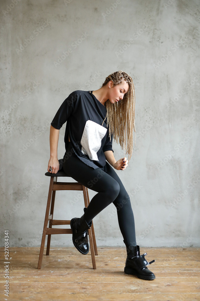 Full-size photo of a young woman with dreadlocks wearing oversize shirt sitting sits on the bar chair and posing against stone background. Full-size photo of a young woman with dreadlocks wearing oversize shirt sitting sits on the bar chair and posing against stone background.