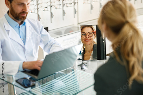Obraz na plátně Woman looks in the mirror trying to choose glasses on the advice of an ophthalmo