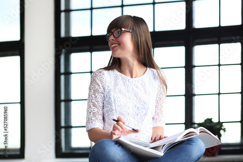 Young, beautiful, cute girl with bangs in glasses sitting on the windowsil and studying.