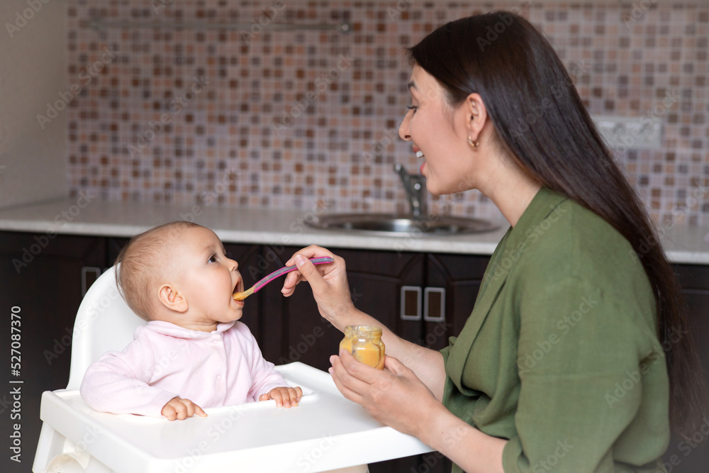 Baby food. A baby in a high chair eats vegetable puree from a spoon. Mom feeds the baby from a jar. Kitchen. Lifestyle. Baby food. A baby in a high chair eats vegetable puree from a spoon. Mom feeds the baby from a jar. Kitchen. Lifestyle.