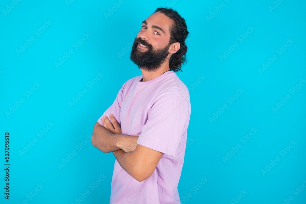 Portrait of young bearded man wearing violet T-shirt over blue studio background standing with folded arms and smiling Portrait of young bearded man wearing violet T-shirt over blue studio background standing with folded arms and smiling