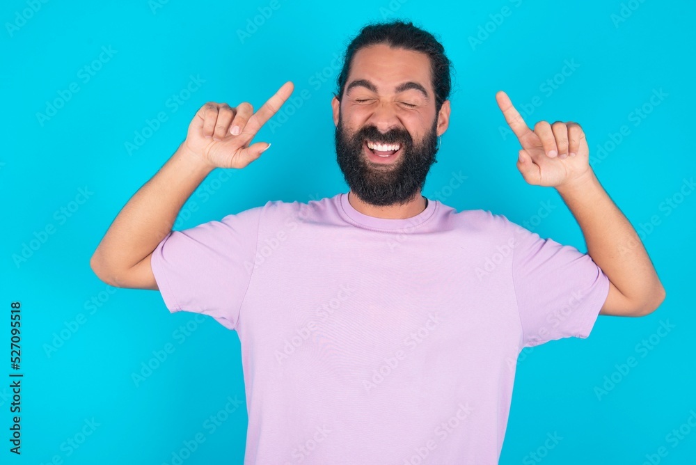 Photo of crazy young bearded man wearing violet T-shirt over blue studio background screaming and pointing with fingers at hair closed eyes Photo of crazy young bearded man wearing violet T-shirt over blue studio background screaming and pointing with fingers at hair closed eyes