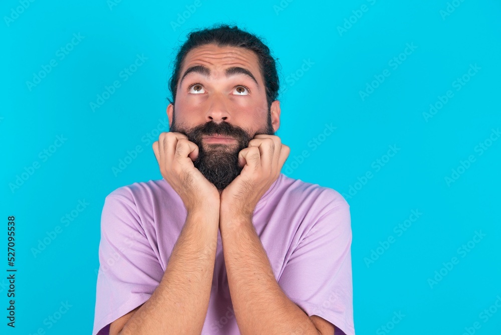 Portrait of sad young bearded man wearing violet T-shirt over blue studio background hands face look empty space