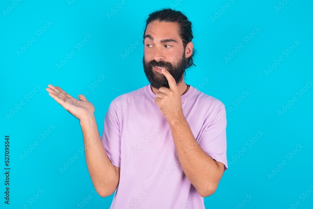 Positive young bearded man wearing violet T-shirt over blue studio background advert promo touches teeth with finger. Positive young bearded man wearing violet T-shirt over blue studio background advert promo touches teeth with finger.