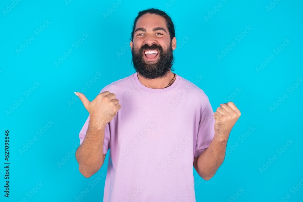 Hooray cool young bearded man wearing violet T-shirt over blue studio background point back empty space hand fist Hooray cool young bearded man wearing violet T-shirt over blue studio background point back empty space hand fist