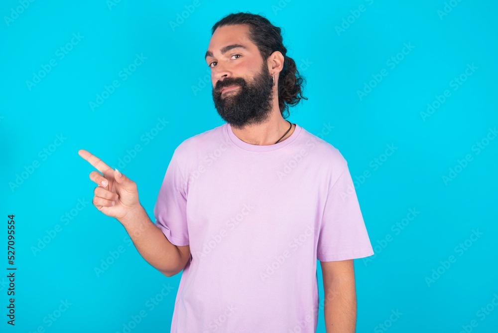 young bearded man wearing violet T-shirt over blue studio background points to side on blank space demonstrates advertisement. People and promotion concept young bearded man wearing violet T-shirt over blue studio background points to side on blank space demonstrates advertisement. People and promotion concept