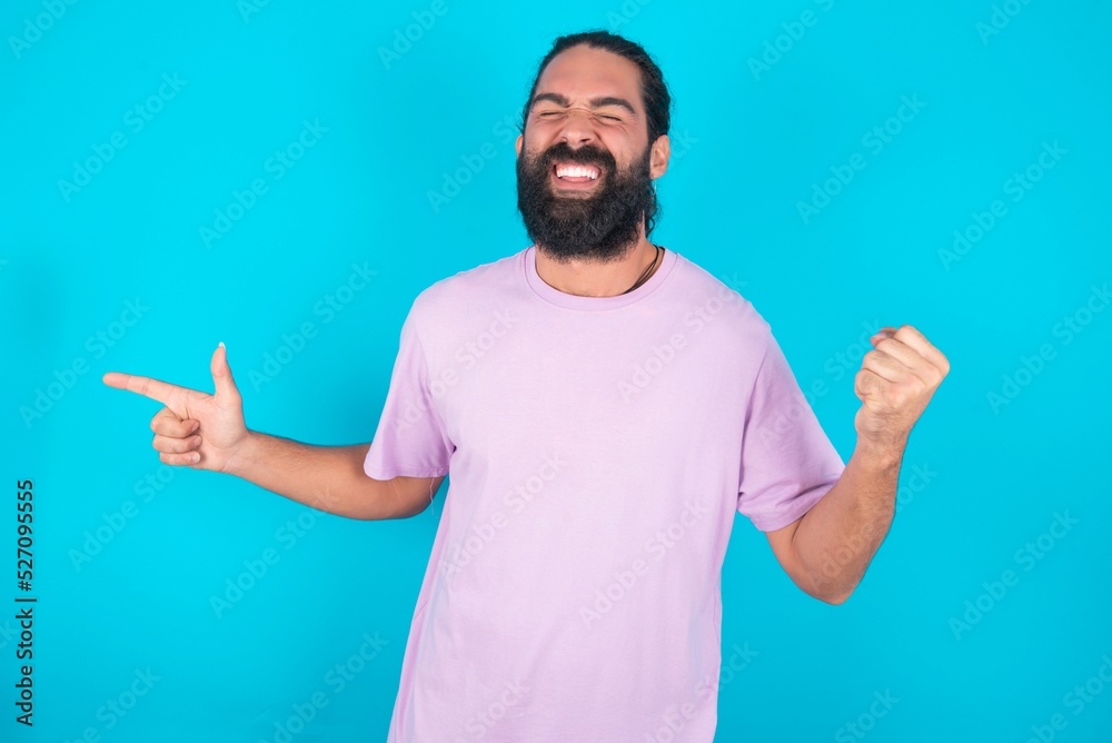 Cheerful young bearded man wearing violet T-shirt over blue studio background showing copy space ad celebrating luck Cheerful young bearded man wearing violet T-shirt over blue studio background showing copy space ad celebrating luck
