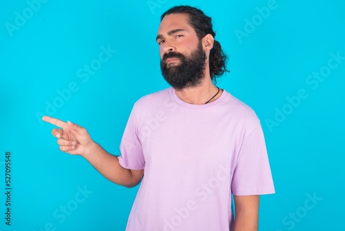 Serious young bearded man wearing violet T-shirt over blue studio background smirks face points away on copy space shows something unpleasant. Look at this advertisement. Big price concept © Jihan