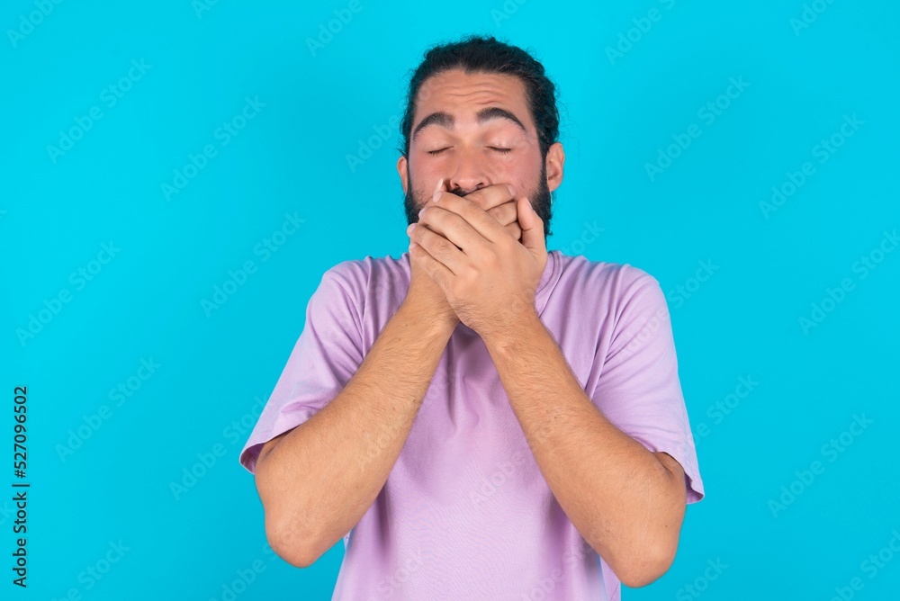 Stunned young bearded man wearing violet T-shirt over blue studio background covers mouth with both hands being afraid from something or after hearing stunning gossips. Stunned young bearded man wearing violet T-shirt over blue studio background covers mouth with both hands being afraid from something or after hearing stunning gossips.