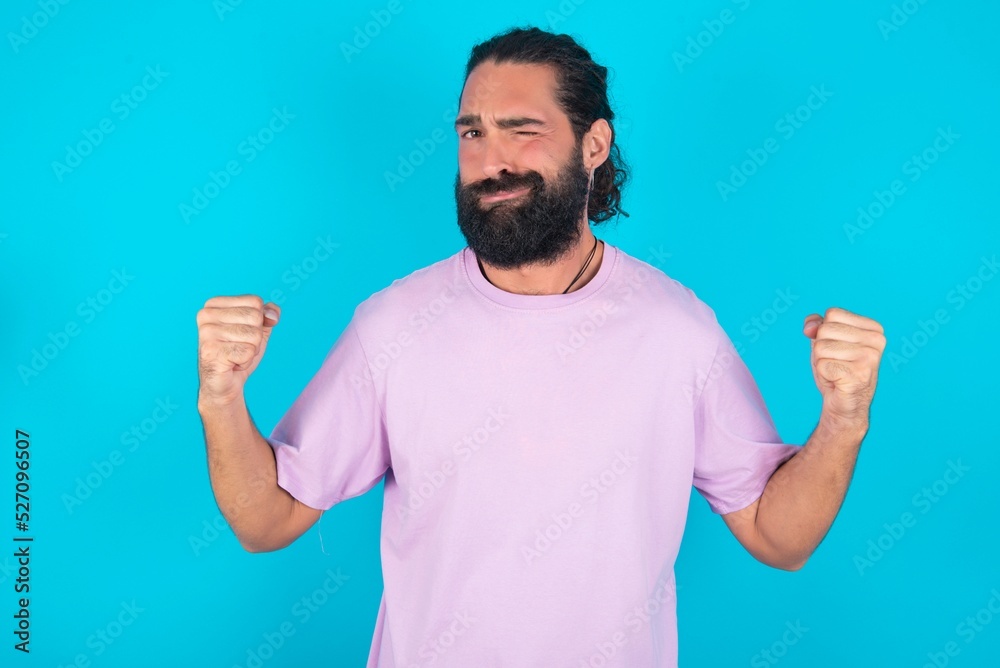 Irritated young bearded man wearing violet T-shirt over blue studio background blows cheeks with anger and raises clenched fists expresses rage and aggressive emotions. Furious model Irritated young bearded man wearing violet T-shirt over blue studio background blows cheeks with anger and raises clenched fists expresses rage and aggressive emotions. Furious model