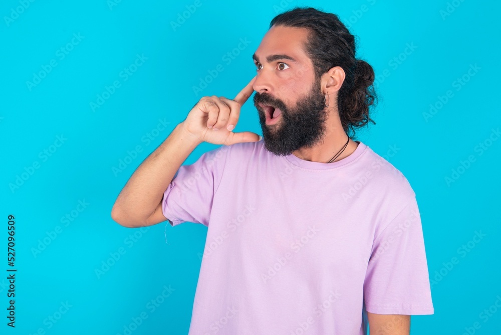 Astonished young bearded man wearing violet T-shirt over blue studio background looks aside surprisingly with opened mouth.