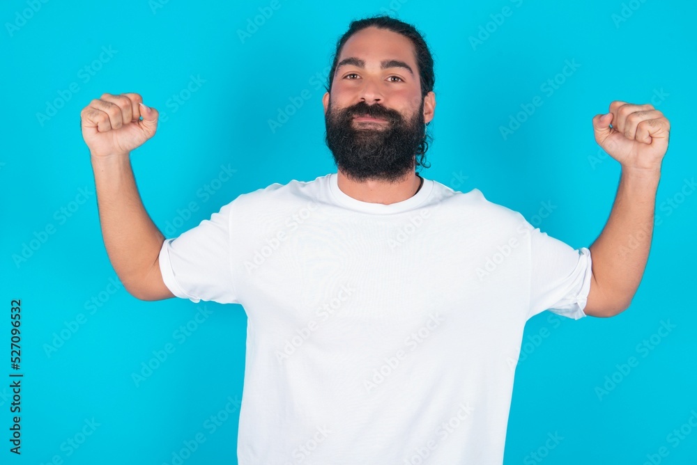 Strong powerful young bearded man wearing white T-shirt over blue studio background toothy smile, raises arms and shows biceps. Look at my muscles! Strong powerful young bearded man wearing white T-shirt over blue studio background toothy smile, raises arms and shows biceps. Look at my muscles!