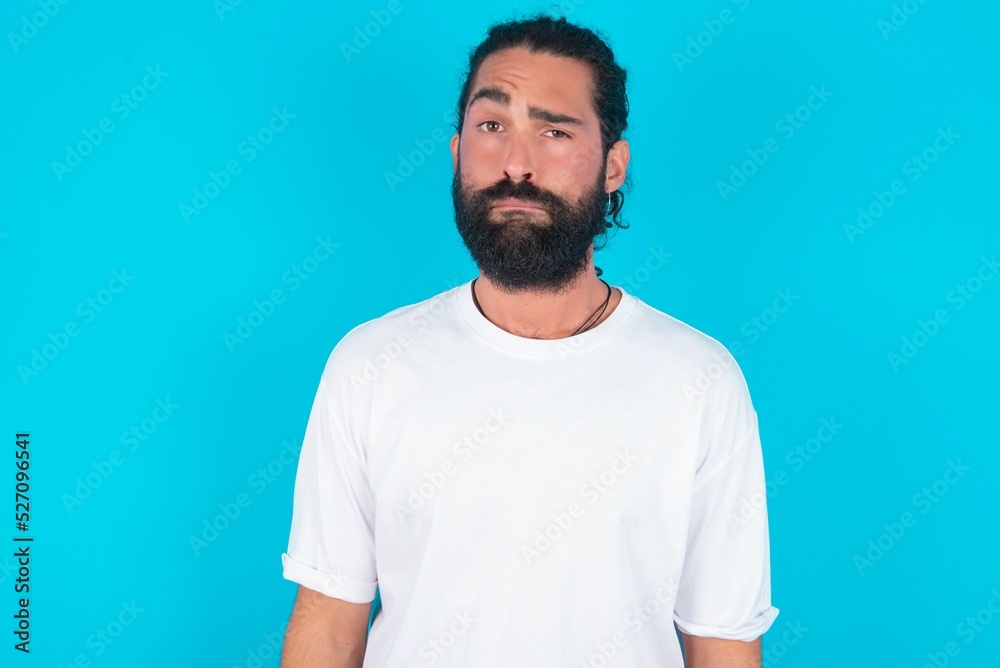 Portrait of dissatisfied young bearded man wearing white T-shirt over blue studio background smirks face, purses lips and looks with annoyance at camera, discontent hearing something unpleasant Portrait of dissatisfied young bearded man wearing white T-shirt over blue studio background smirks face, purses lips and looks with annoyance at camera, discontent hearing something unpleasant