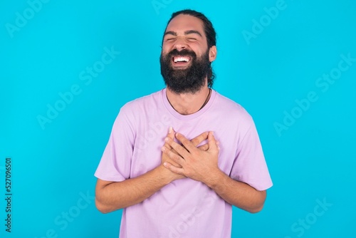 young bearded man wearing violet T-shirt over blue studio background expresses happiness, laughs pleasantly, keeps hands on heart