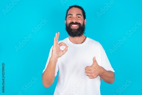 young bearded man wearing white T-shirt over blue studio background smiling and looking happy, carefree and positive, gesturing victory or peace with one hand