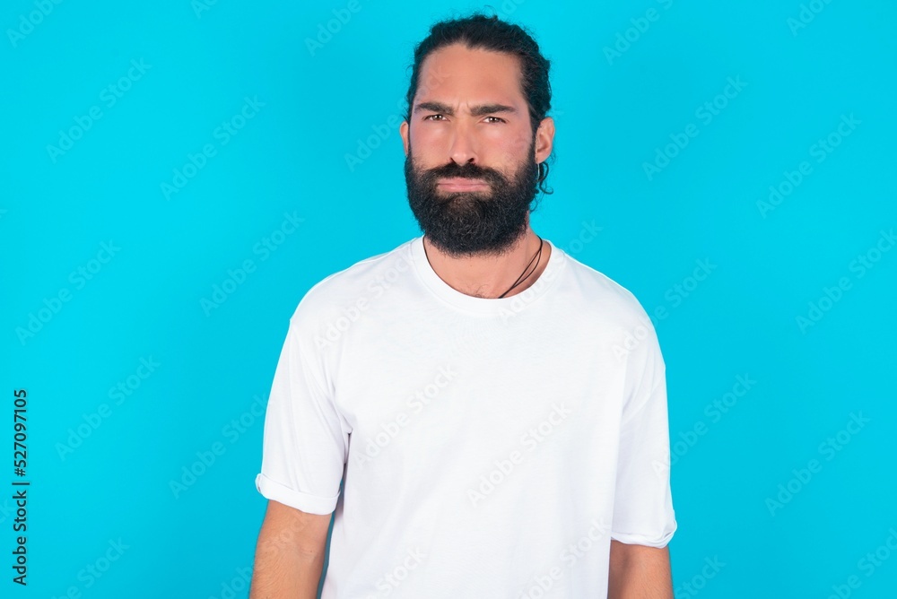 Offended dissatisfied young bearded man wearing white T-shirt over blue studio background with moody displeased expression at camera being disappointed by something Offended dissatisfied young bearded man wearing white T-shirt over blue studio background with moody displeased expression at camera being disappointed by something