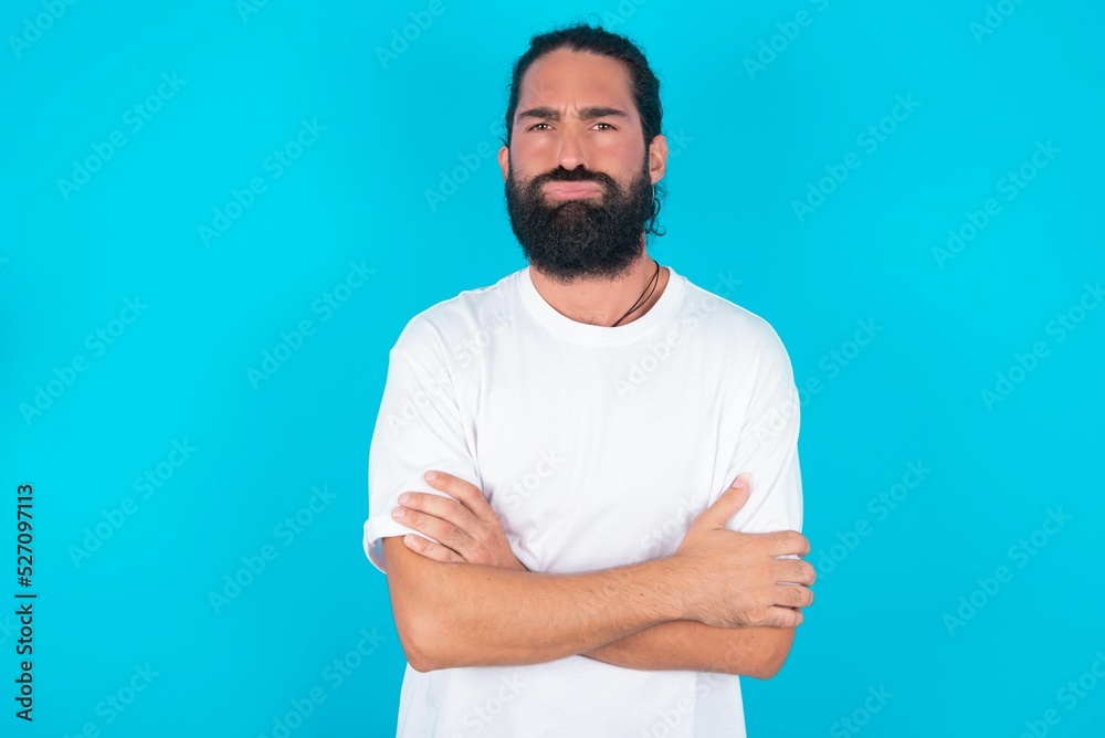 Gloomy dissatisfied young bearded man wearing white T-shirt over blue studio background looks with miserable expression at camera from under forehead, makes unhappy grimace Gloomy dissatisfied young bearded man wearing white T-shirt over blue studio background looks with miserable expression at camera from under forehead, makes unhappy grimace