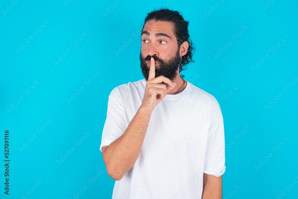 young bearded man wearing white T-shirt over blue studio background makes silence gesture, keeps index finger to lips makes hush sign. Asks not to share secret. young bearded man wearing white T-shirt over blue studio background makes silence gesture, keeps index finger to lips makes hush sign. Asks not to share secret.