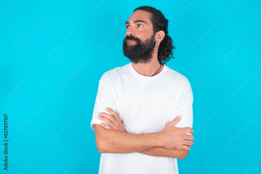 Charming thoughtful young bearded man wearing white T-shirt over blue studio background stands with arms folded concentrated somewhere with pensive expression thinks what to do Charming thoughtful young bearded man wearing white T-shirt over blue studio background stands with arms folded concentrated somewhere with pensive expression thinks what to do