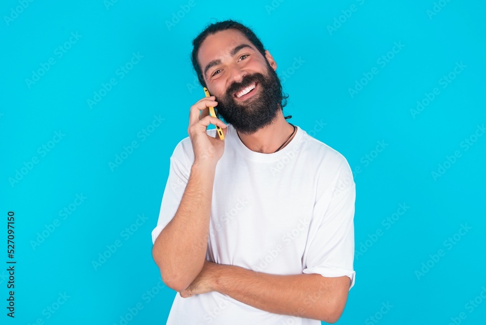 Portrait of a smiling young bearded man wearing white T-shirt over blue studio background talking on mobile phone. Business, confidence and communication concept. Portrait of a smiling young bearded man wearing white T-shirt over blue studio background talking on mobile phone. Business, confidence and communication concept.