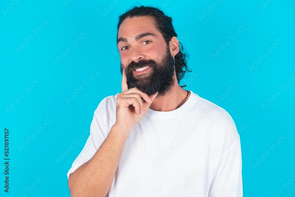 Carefree successful young bearded man wearing white T-shirt over blue studio background touching jawline gazing camera tilting head grinning white teeth delighted. Dental care concept. Carefree successful young bearded man wearing white T-shirt over blue studio background touching jawline gazing camera tilting head grinning white teeth delighted. Dental care concept.
